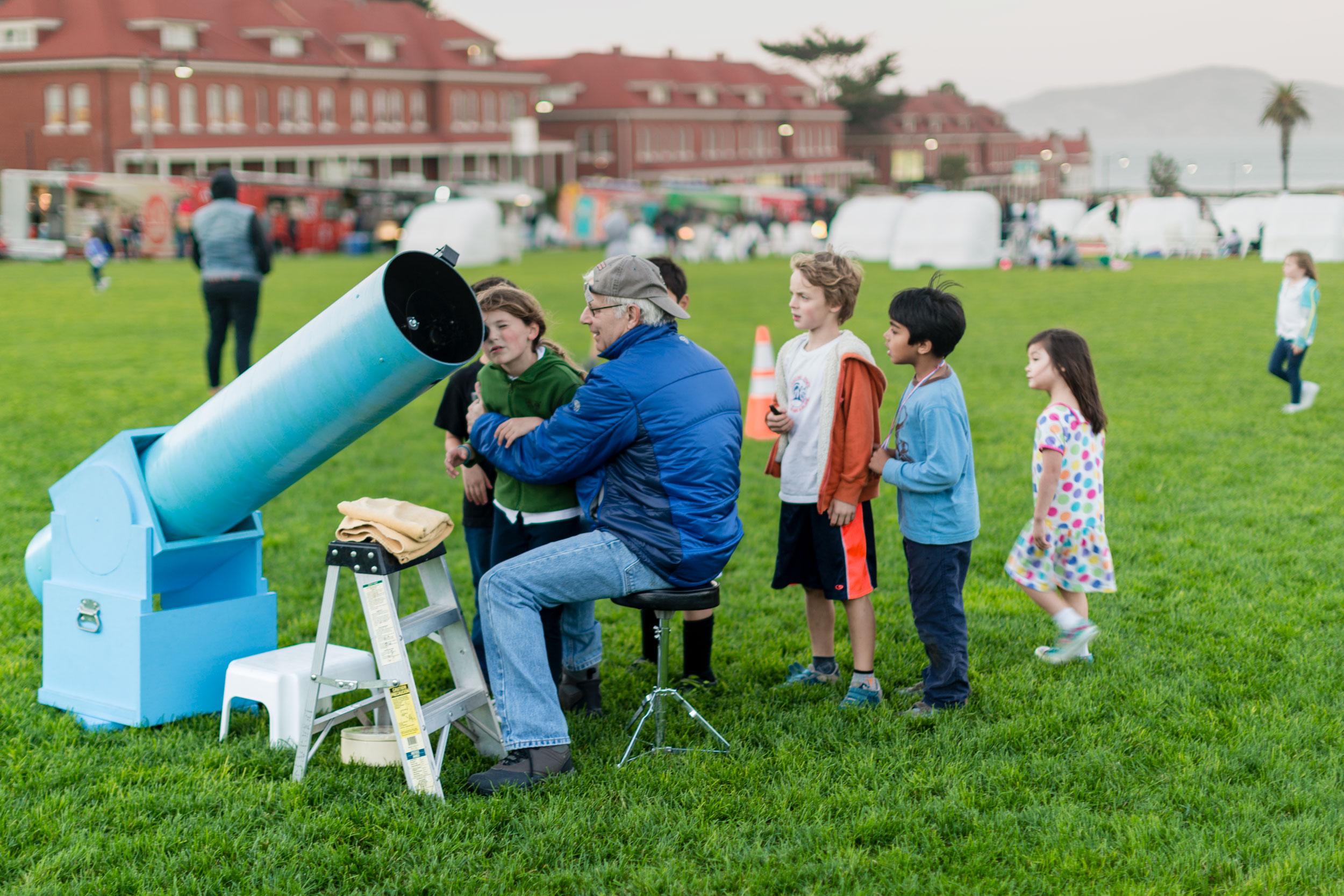 Man seated and showing line of children how to use telescope