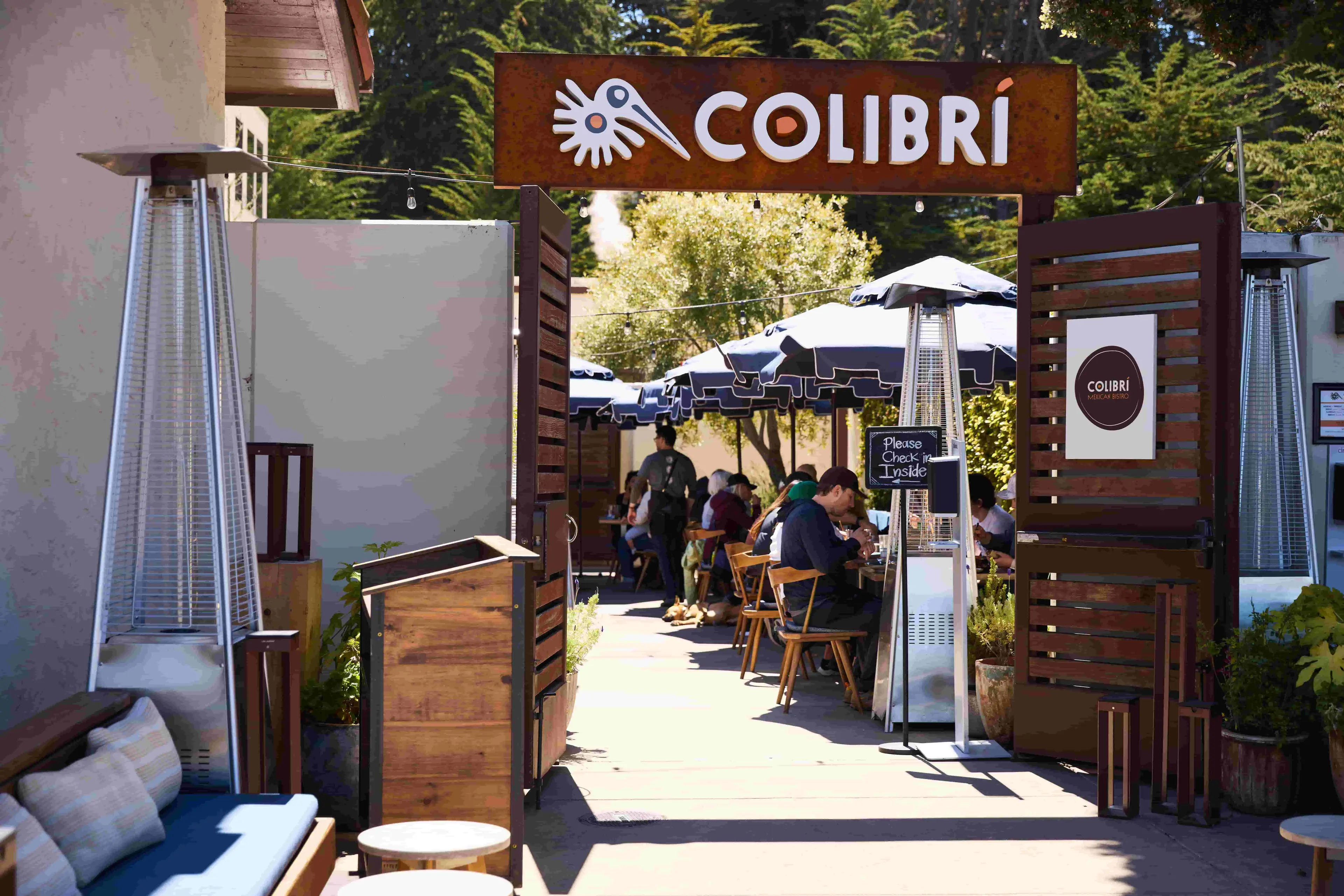 Entrance to restaurant with a hostess stand and heater in the foreground and people seated, eating inside the restaurant