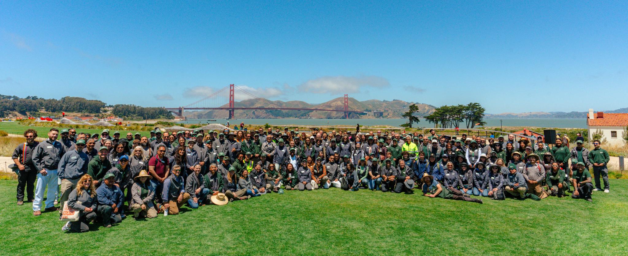 Group photo of Presidio Trust employees standing on a lawn with Golden Gate Bridge in the background.