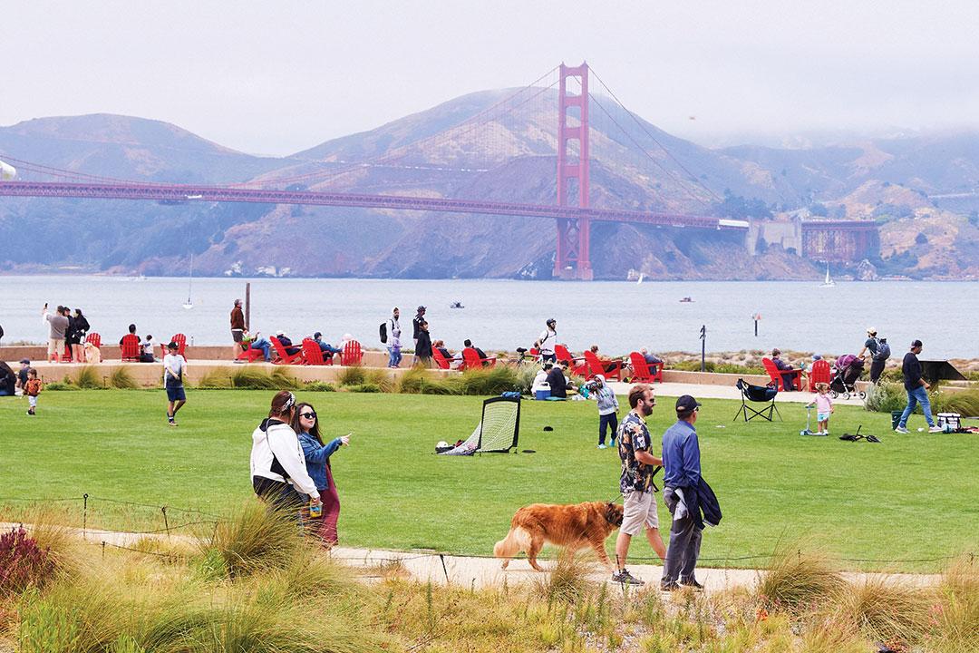 Two men and a dog walking in front of two women talking at the Presidio Tunnel Tops with the Golden Gate Bridge in the background.