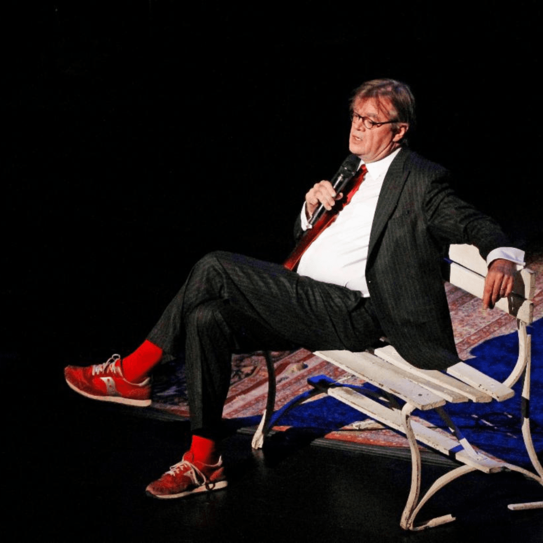 Garrision Keilor sitting on a bench on a stage with a microphone