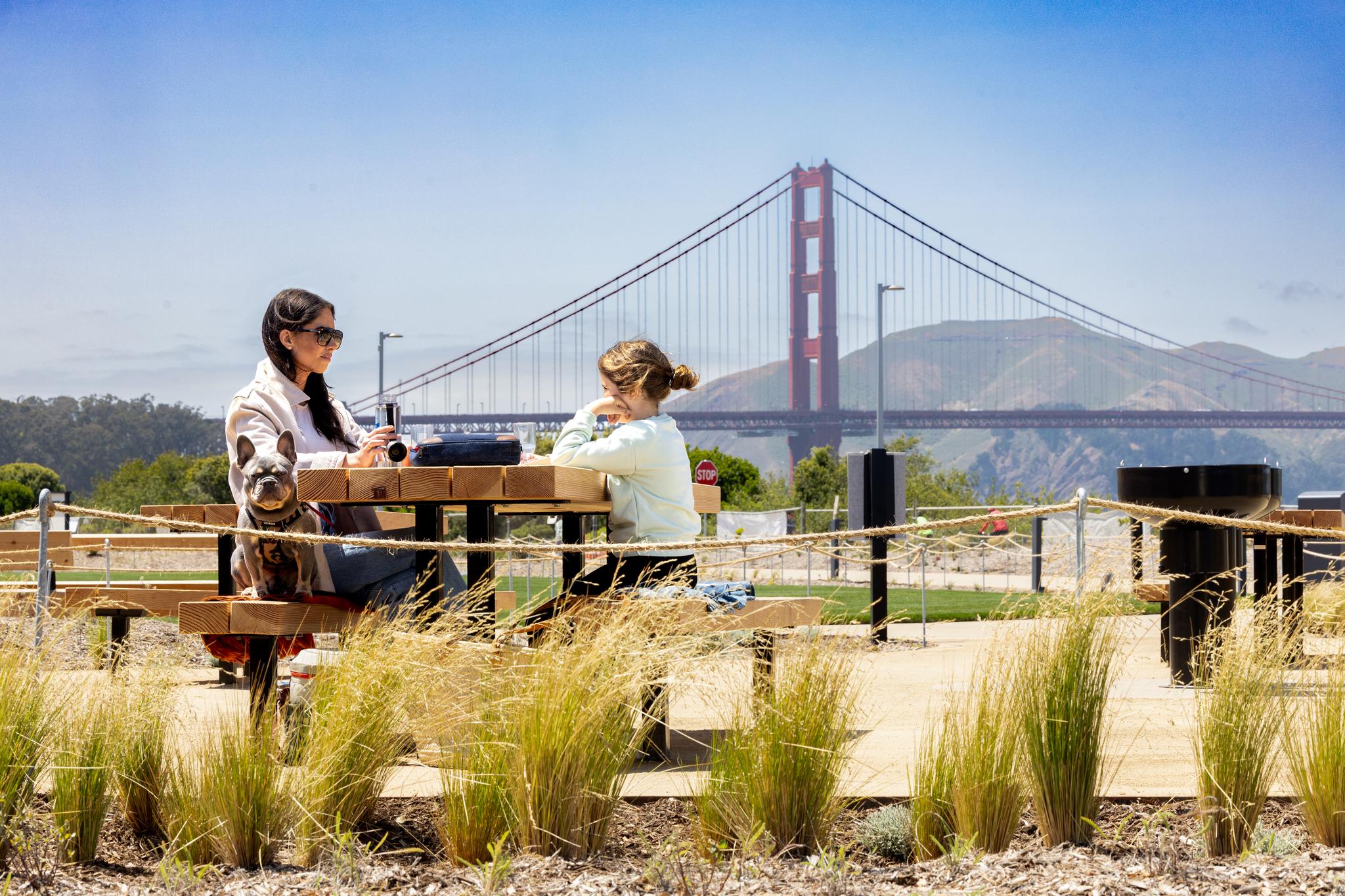Woman, girl, and dog sitting on a picnic bench at Outpost Meadow, with Golden Gate Bridge background