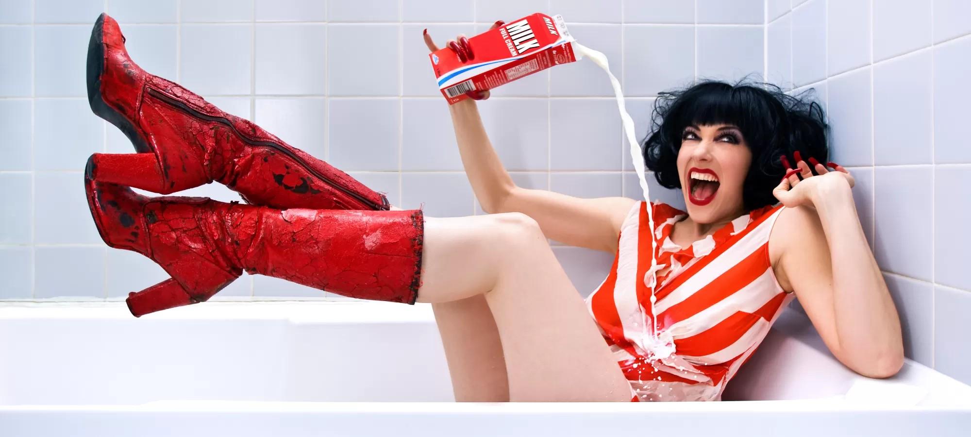 Woman in red and white striped outfit and red boots pouring milk on herself in a bathtub