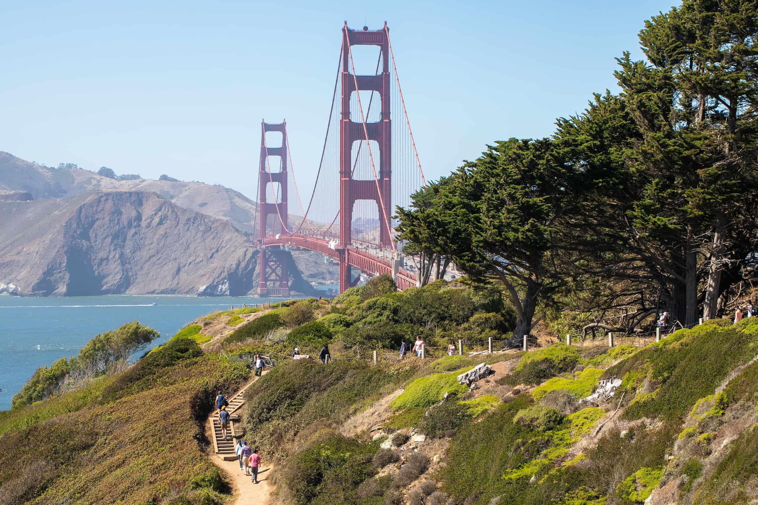 A distant shot of people walking along the Batteries to Bluffs trail with the Golden Gate Bridge in the background on a sunny day.