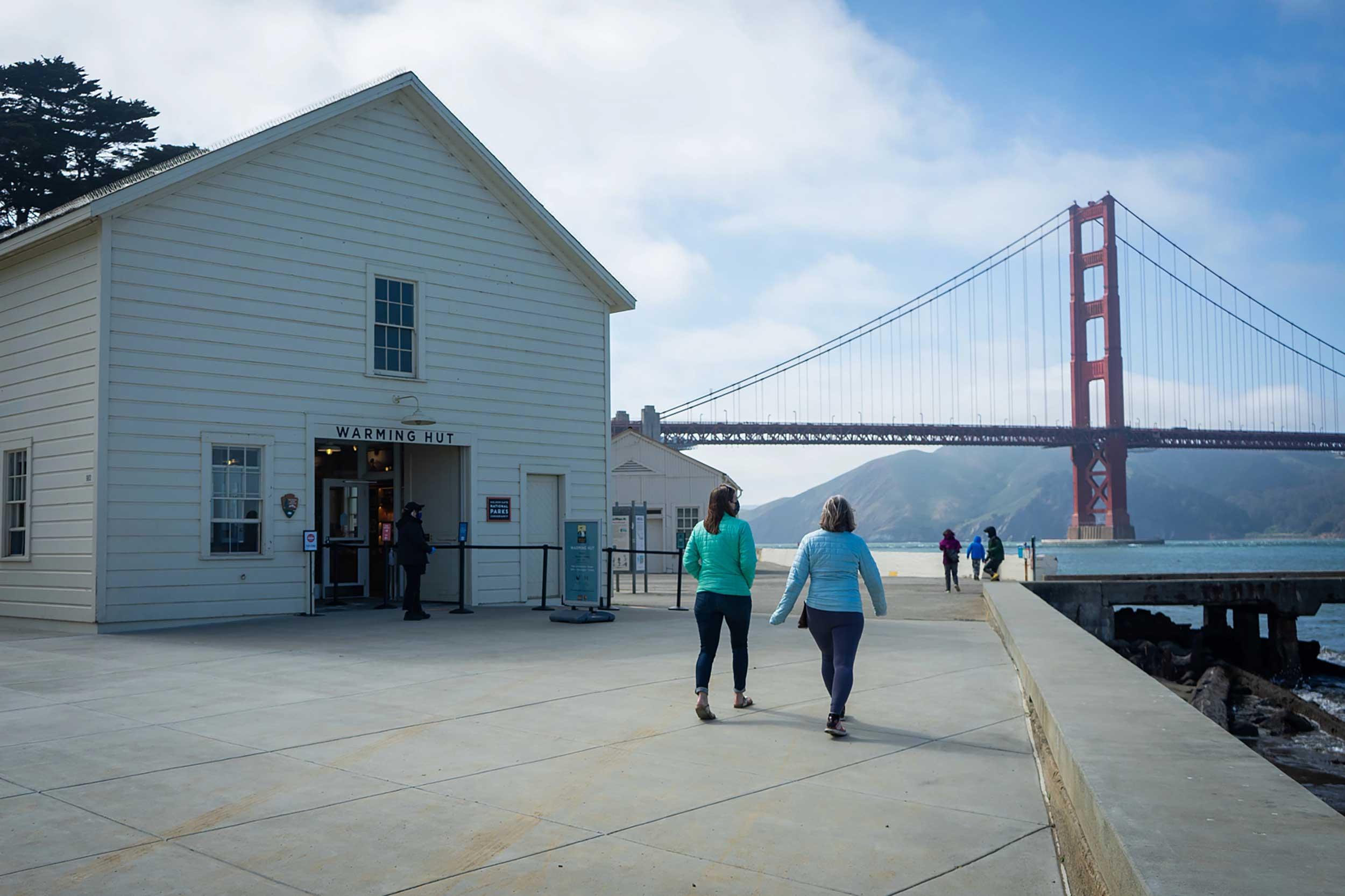 Two women walking in front of the Warming Hut Park Store at the Presidio of San Francisco with the Golden Gate Bridge in the background. Photo by Ryan Curran White.