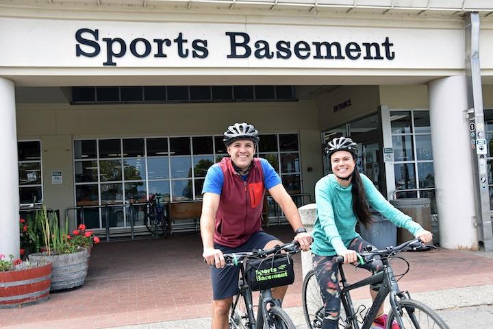 A man and woman resting on bicycles in front of Sports Basement.