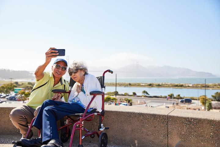 A man and a woman sitting in a wheelchair take a photo from a scenic overlook. Photo by Rachel Styer.