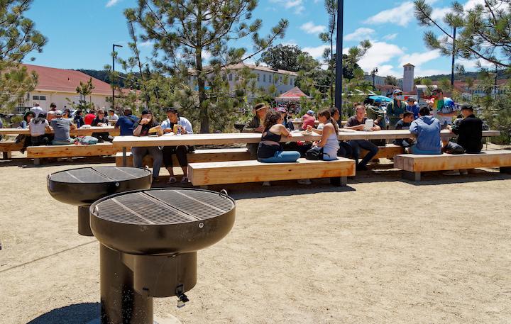 Many visitors sitting at a picnic table with companion seating, with a grill in the foreground. Photo by Dan Friedman.