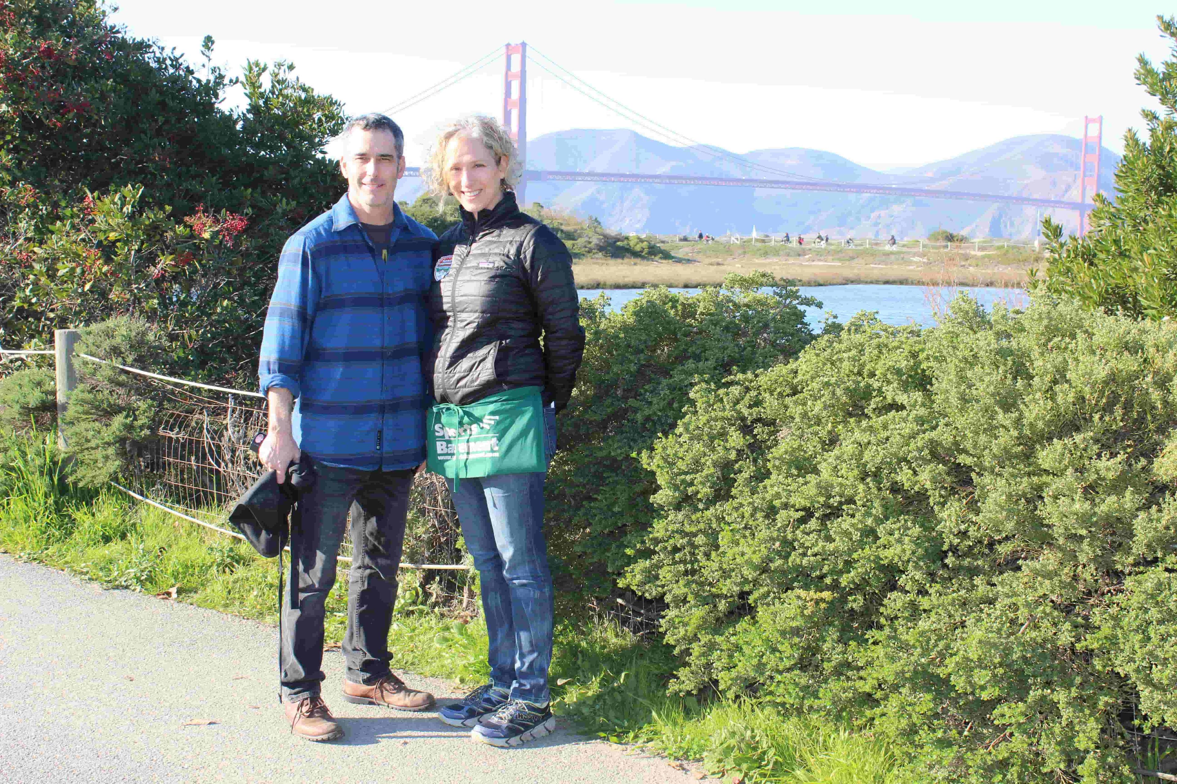 Two people standing near Crissy Marsh with Golden Gate Bridge in background.