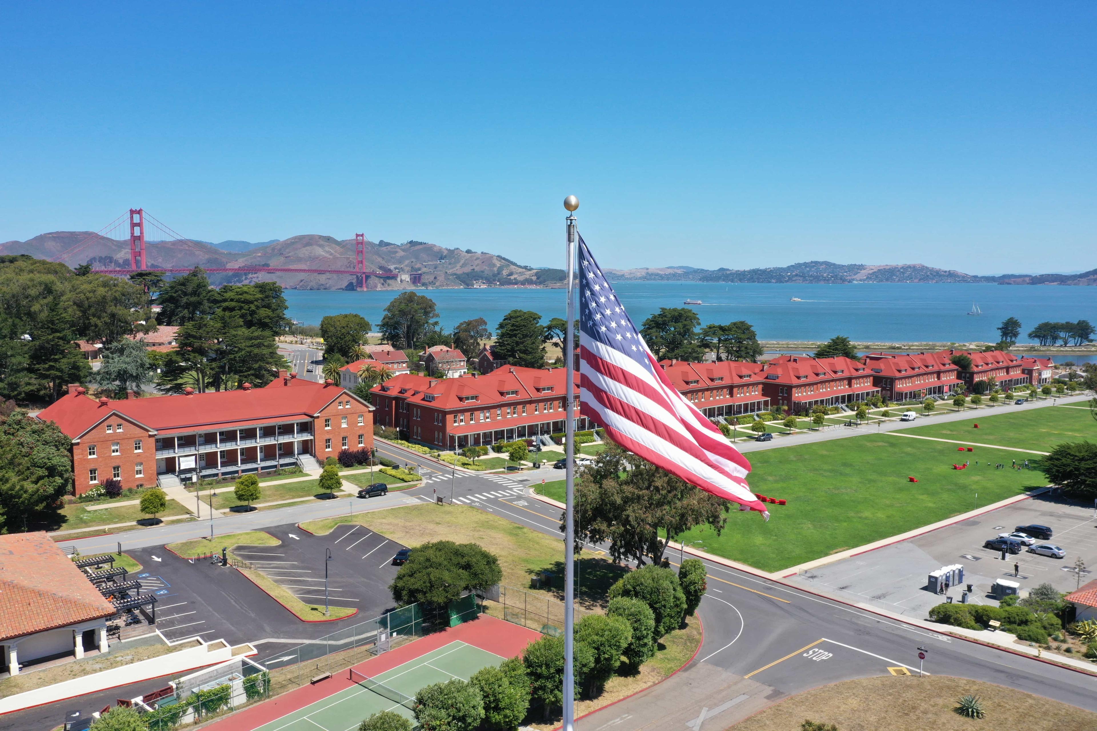 Image of American flag waving in foreground with historic barracks, the Main Parade Ground of the Presidio and the Golden Gate Bridge in the background.