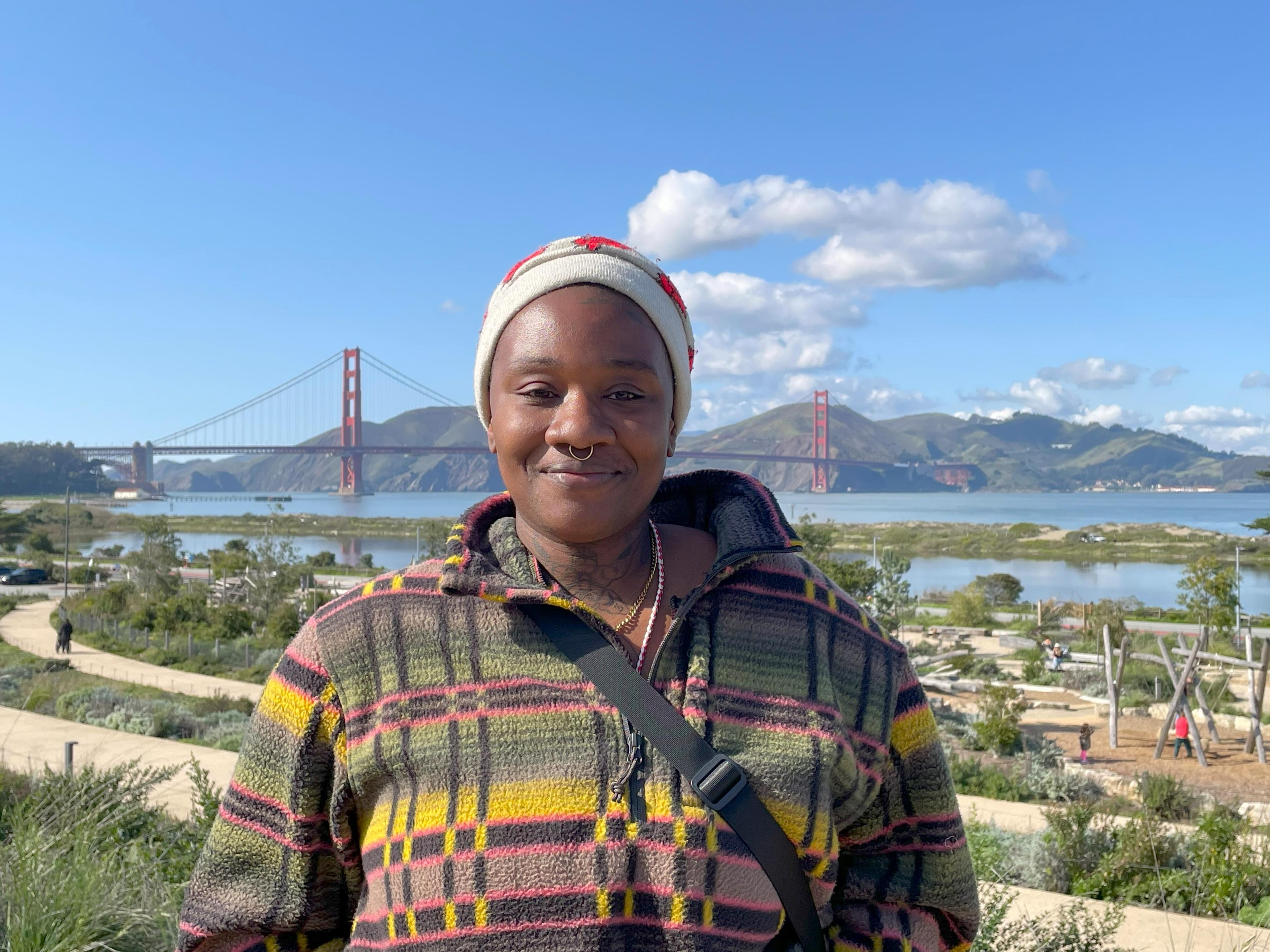 Image of a woman standing and smiling in a plaid jacket on the Presidio Tunnel Tops with the Golden Gate Bridge in the background.