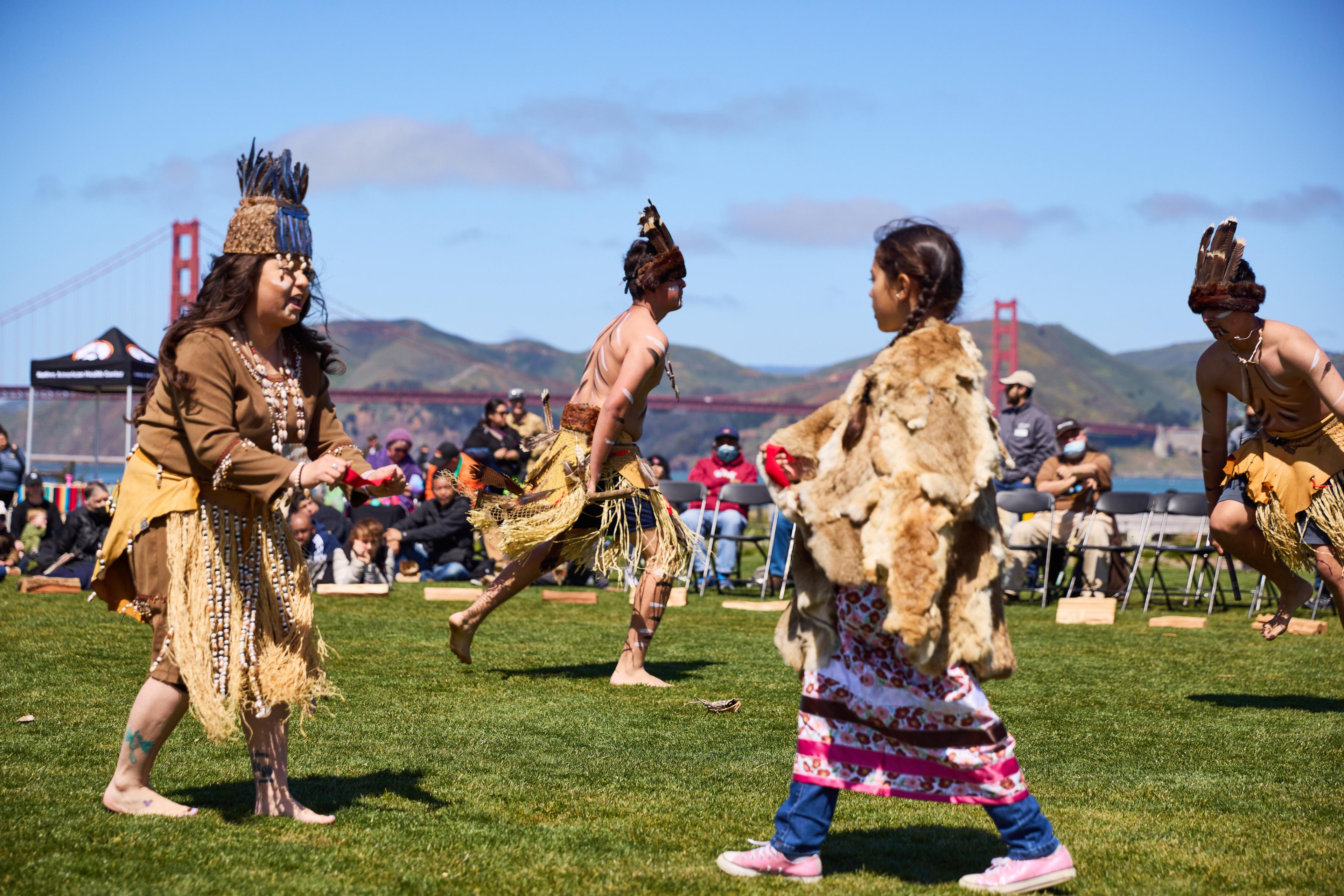 Group of individuals dressed in indigenous attire dancing with the Golden Gate Bridge in the backgrounf=d.